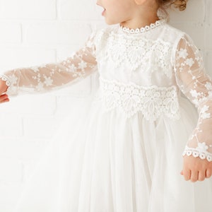 May include: A white tulle dress with long sleeves and lace detailing. The dress has a high neckline and a full skirt.