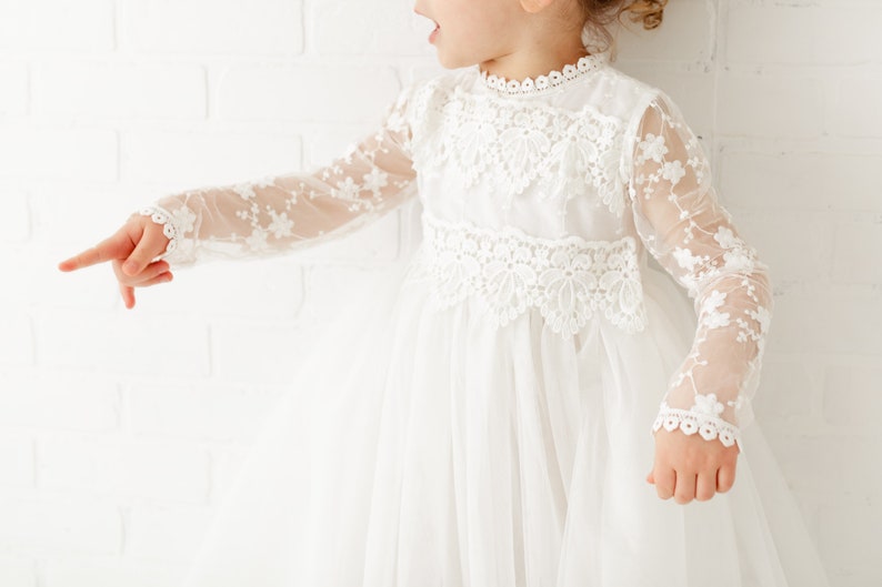 May include: A white tulle dress with long sleeves and lace detailing. The dress has a high neckline and a full skirt.