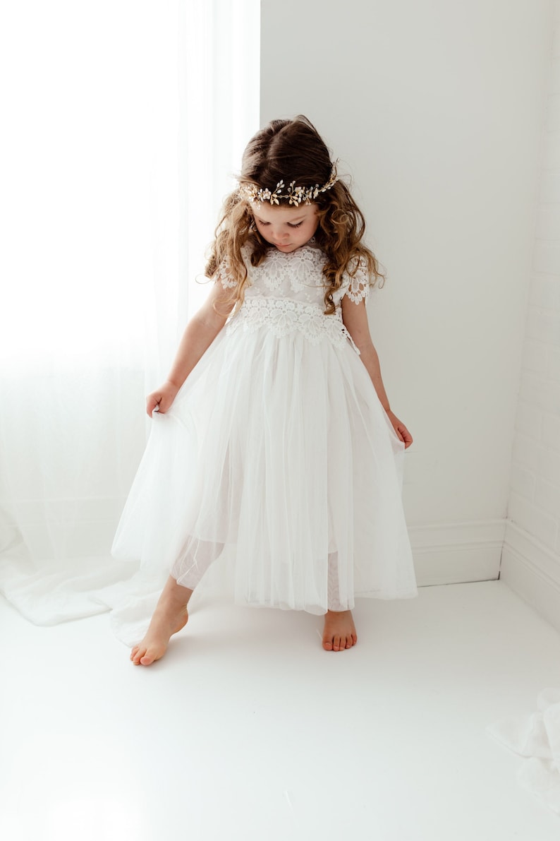 May include: A young girl wearing a white tulle dress with lace detailing and a gold leaf crown. The dress has a full skirt and short sleeves. The girl is standing on a white background.