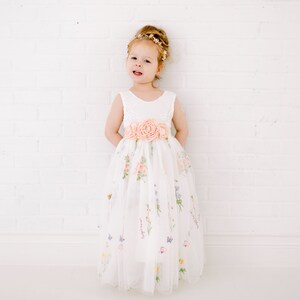 Puede incluir: Una ni&ntilde;a peque&ntilde;a con un vestido de tul blanco con estampado floral y una faja de flores de melocot&oacute;n. Lleva una diadema dorada y tiene el pelo recogido hacia atr&aacute;s.