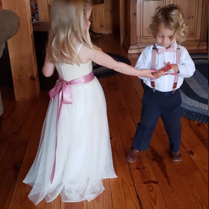 May include: A young girl wearing a white tulle dress with a pink sash and a young boy in a white shirt, navy blue trousers, and brown braces. The boy is holding a small red gift box. They are standing on a wooden floor.