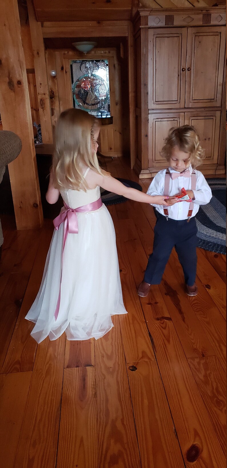 May include: A young girl wearing a white tulle dress with a pink sash and a young boy in a white shirt, navy blue trousers, and brown braces. The boy is holding a small red gift box. They are standing on a wooden floor.