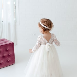 Pu&ograve; includere: Una bambina che indossa un abito bianco di tulle con maniche di pizzo e un cerchietto bianco. L'abito ha una schiena di pizzo e un fiocco di raso.