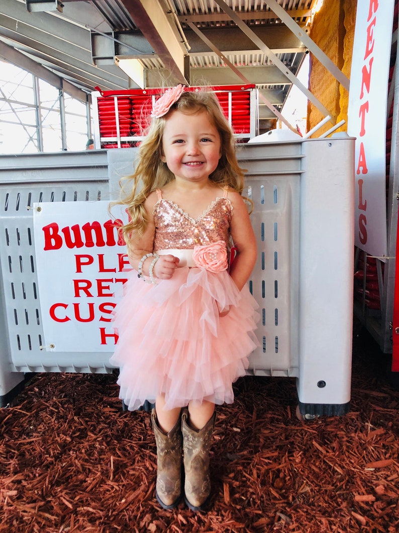 May include: A young girl wearing a pink tulle dress with a sequined bodice and a rose-colored flower on the waist. She is wearing brown cowboy boots and a pink flower headband. The girl is standing in front of a gray storage container.