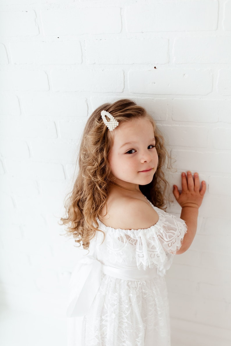 May include: A young girl with curly blonde hair wearing a white off-the-shoulder lace dress with a satin sash. She is wearing a pearl hair clip and has a sweet expression on her face.
