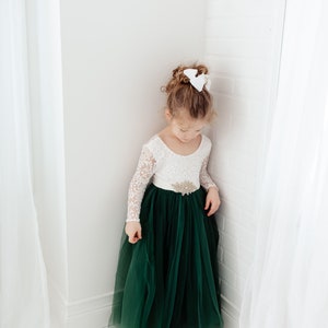 K&ouml;nnte beinhalten: Ein junges M&auml;dchen tr&auml;gt ein wei&szlig;es, lang&auml;rmliges Kleid aus Spitze mit einem gr&uuml;nen T&uuml;llrock und einer wei&szlig;en Schleife im Haar.