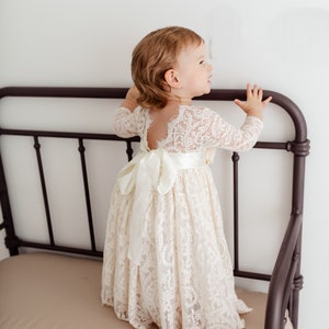 May include: A young girl wearing a white lace dress with a satin ribbon tied at the waist. She is standing on a brown bench with her hands on the back.