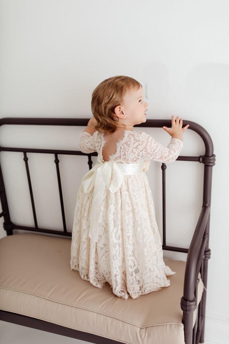 May include: A young girl wearing a white lace dress with a satin ribbon tied at the waist. She is standing on a brown bench with her hands on the back.