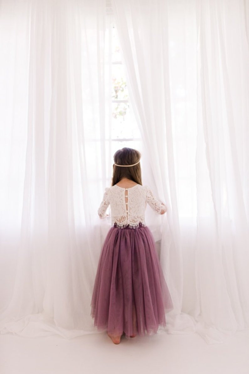 May include: A young girl wearing a white lace top with a button-up back and a long, dusty rose tulle skirt. She is standing in front of a window with white curtains.