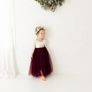 May include: A young girl wearing a white long-sleeved dress with a burgundy tulle skirt and a flower crown. She is standing in front of a white brick wall.