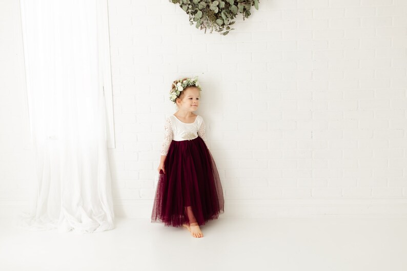 May include: A young girl wearing a white long-sleeved dress with a burgundy tulle skirt and a flower crown. She is standing in front of a white brick wall.