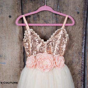 May include: A tulle dress with a rose gold sequin bodice and a pink flower sash. The dress is on a pink polka dot hanger against a wooden background.