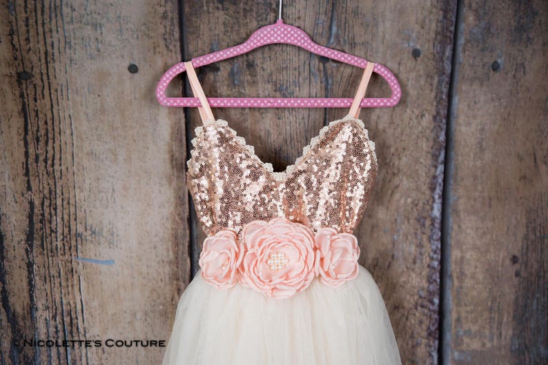 May include: A tulle dress with a rose gold sequin bodice and a pink flower sash. The dress is on a pink polka dot hanger against a wooden background.