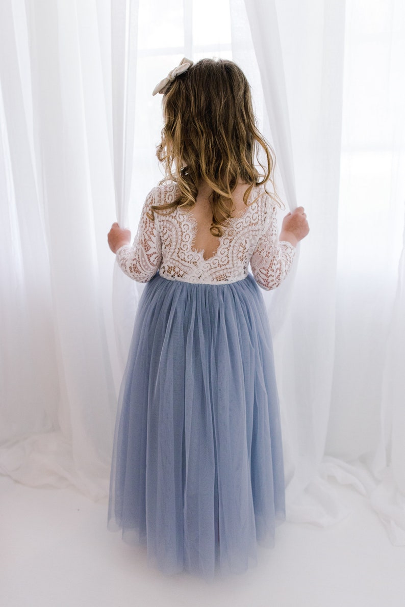May include: A young girl wearing a white lace bodice dress with a light blue tulle skirt. She is standing in front of a white curtain and looking out a window.