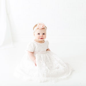 May include: A white lace dress with short sleeves and a full skirt, featuring a delicate floral pattern throughout the fabric. The dress is paired with a matching white headband adorned with a large, soft peach-colored fabric flower accessory.