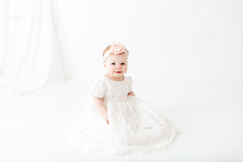 May include: A white lace dress with short sleeves and a full skirt, featuring a delicate floral pattern throughout the fabric. The dress is paired with a matching white headband adorned with a large, soft peach-colored fabric flower accessory.