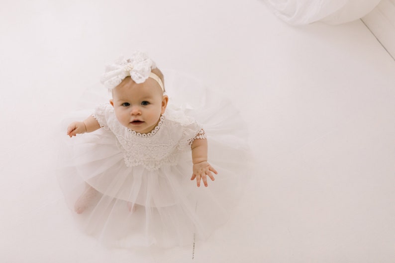 May include: A white infant dress featuring a lace bodice with short sleeves and a multi-layered tulle skirt. The garment is paired with a matching white lace headband adorned with a large bow, creating a coordinated ensemble for a young child.