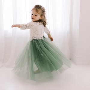 May include: A young girl wearing a white lace top and a sage green tulle skirt. She is twirling and smiling.