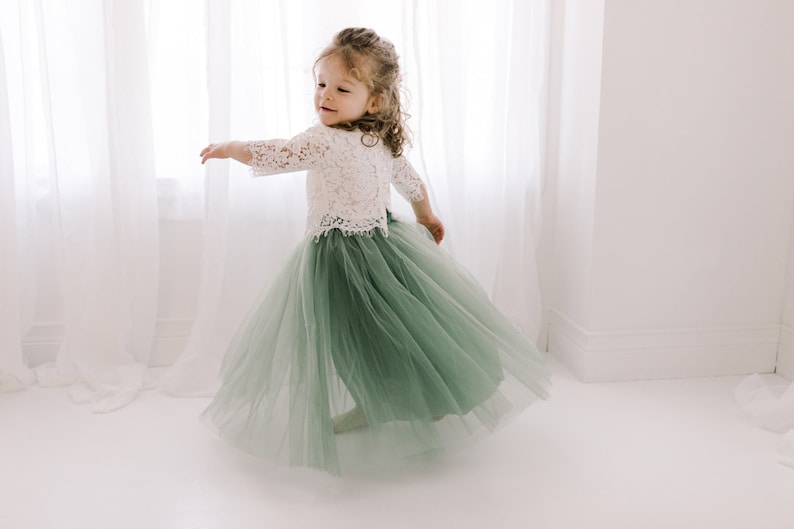May include: A young girl wearing a white lace top and a sage green tulle skirt. She is twirling and smiling.