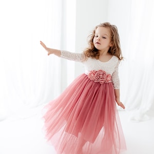 May include: A young girl wearing a white lace top with a pink tulle skirt and a floral sash. The skirt is flowing and the girl is smiling.