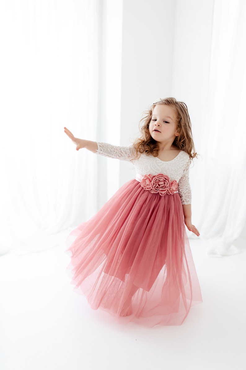 May include: A young girl wearing a white lace top with a pink tulle skirt and a floral sash. The skirt is flowing and the girl is smiling.