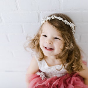 May include: A young girl with long, curly brown hair is wearing a white dress with a pink tulle skirt and a pearl headband. She is smiling and looking up at the camera.