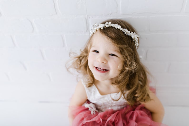 May include: A young girl with long, curly brown hair is wearing a white dress with a pink tulle skirt and a pearl headband. She is smiling and looking up at the camera.