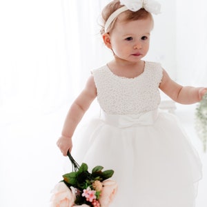 Puede incluir: Una ni&ntilde;a peque&ntilde;a con un vestido blanco de encaje y una diadema blanca con lazo sostiene un ramo de rosas rosas y hojas verdes.