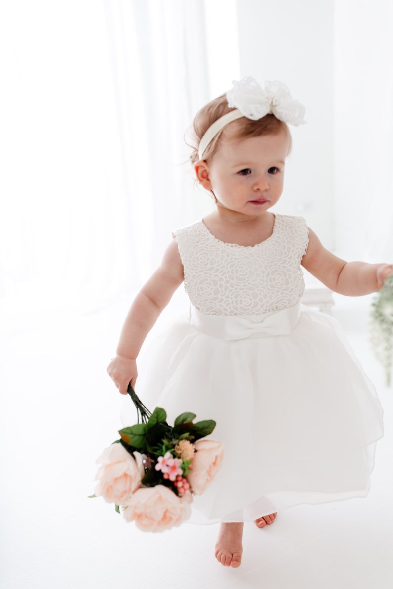 Puede incluir: Una ni&ntilde;a peque&ntilde;a con un vestido blanco de encaje y una diadema blanca con lazo sostiene un ramo de rosas rosas y hojas verdes.