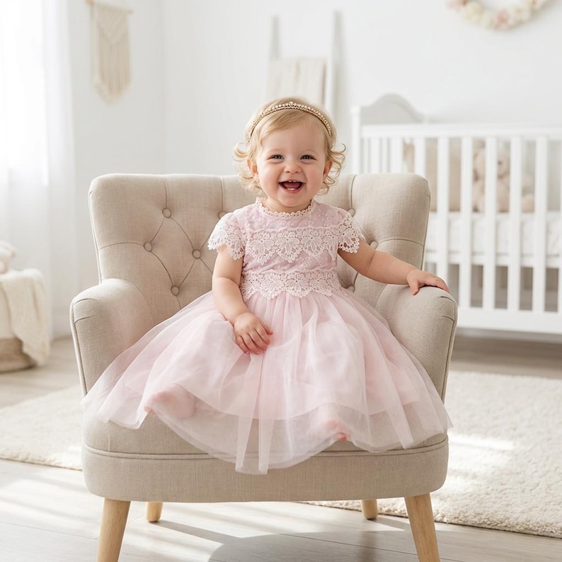 May include: A light pink dress featuring a lace bodice with short sleeves and a layered tulle skirt. The dress is worn with a thin, gold-toned headband adorned with small pearls, creating a soft and delicate appearance for a young child.
