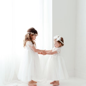 May include: Two young girls in white dresses holding hands. The dresses have lace sleeves and tulle skirts. One girl wears a floral headband, the other a white bow. The setting is a bright, white room with a sheer curtain.