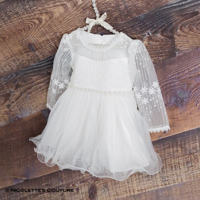 K&ouml;nnte beinhalten: Wei&szlig;es T&uuml;llkleid mit langen &Auml;rmeln, einem perlenbesetzten Ausschnitt und einer perlenbesetzten Taille. Das Kleid hat ein zartes Blumenmuster und einen mehrlagigen Rock.