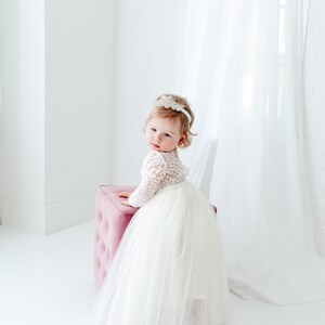 May include: A toddler wearing a white dress with a long-sleeved lace bodice and a full, layered tulle skirt. The dress is paired with a white headband featuring a decorative floral embellishment. The child is positioned next to a pink ottoman.