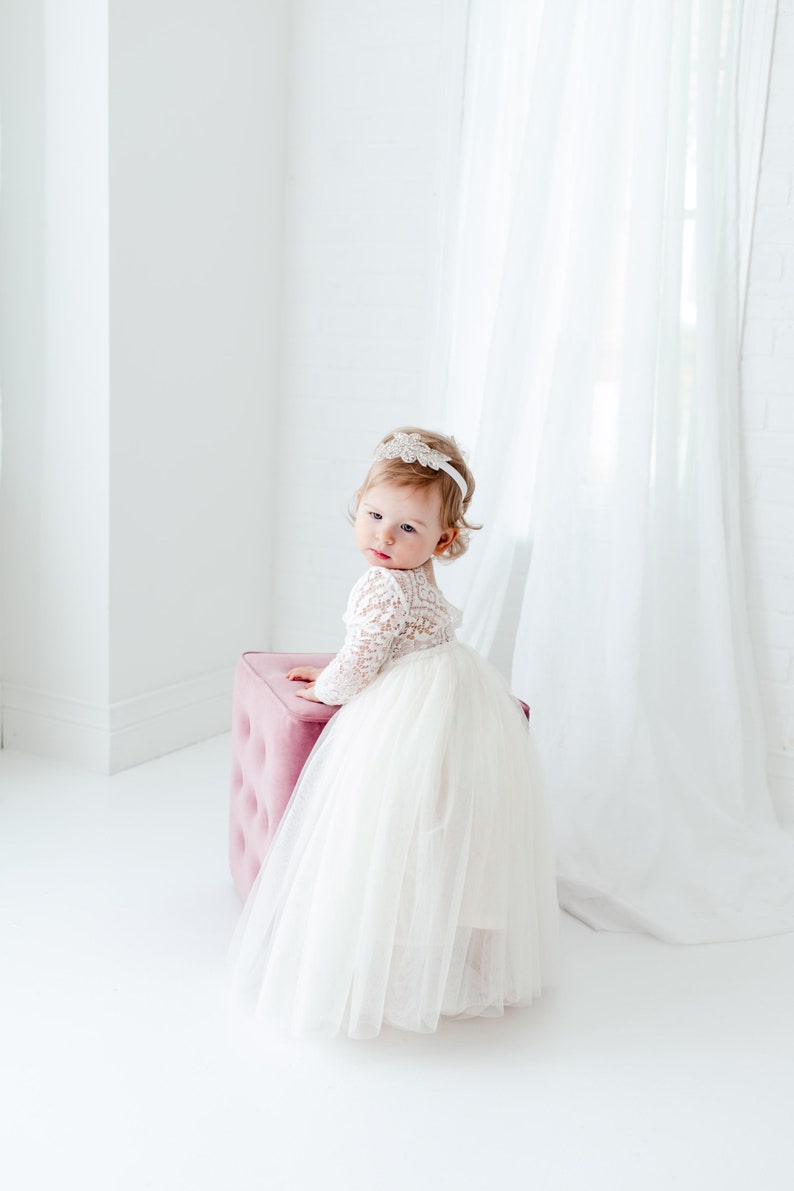 May include: A toddler wearing a white dress with a long-sleeved lace bodice and a full, layered tulle skirt. The dress is paired with a white headband featuring a decorative floral embellishment. The child is positioned next to a pink ottoman.