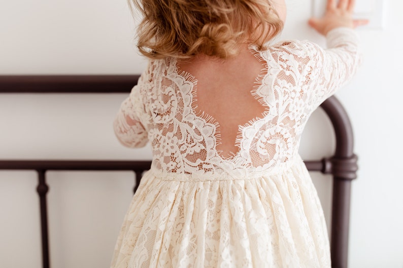 May include: A white lace dress with a low back and long sleeves. The dress is gathered at the waist and has a flowing skirt.