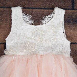 May include: A white lace bodice with pearl embellishments and a pink tulle skirt. The dress has a sleeveless design and a round neckline.