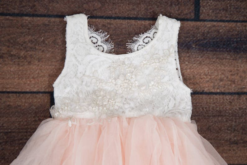 May include: A white lace bodice with pearl embellishments and a pink tulle skirt. The dress has a sleeveless design and a round neckline.