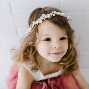 May include: A young girl with long, curly brown hair wearing a white dress with a pink tulle overlay and a pearl and crystal headband.