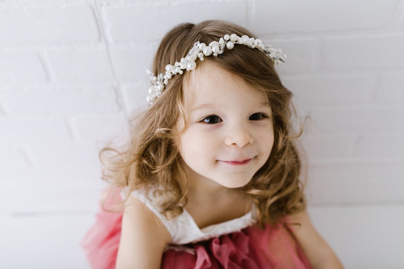 May include: A young girl with long, curly brown hair wearing a white dress with a pink tulle overlay and a pearl and crystal headband.