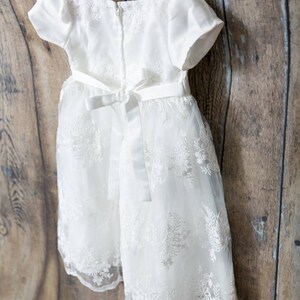 May include: A white dress featuring short puff sleeves, a round neckline, and a back zipper closure. The bodice and skirt are adorned with intricate white floral embroidery over a sheer overlay, finished with a satin ribbon tie at the waist.