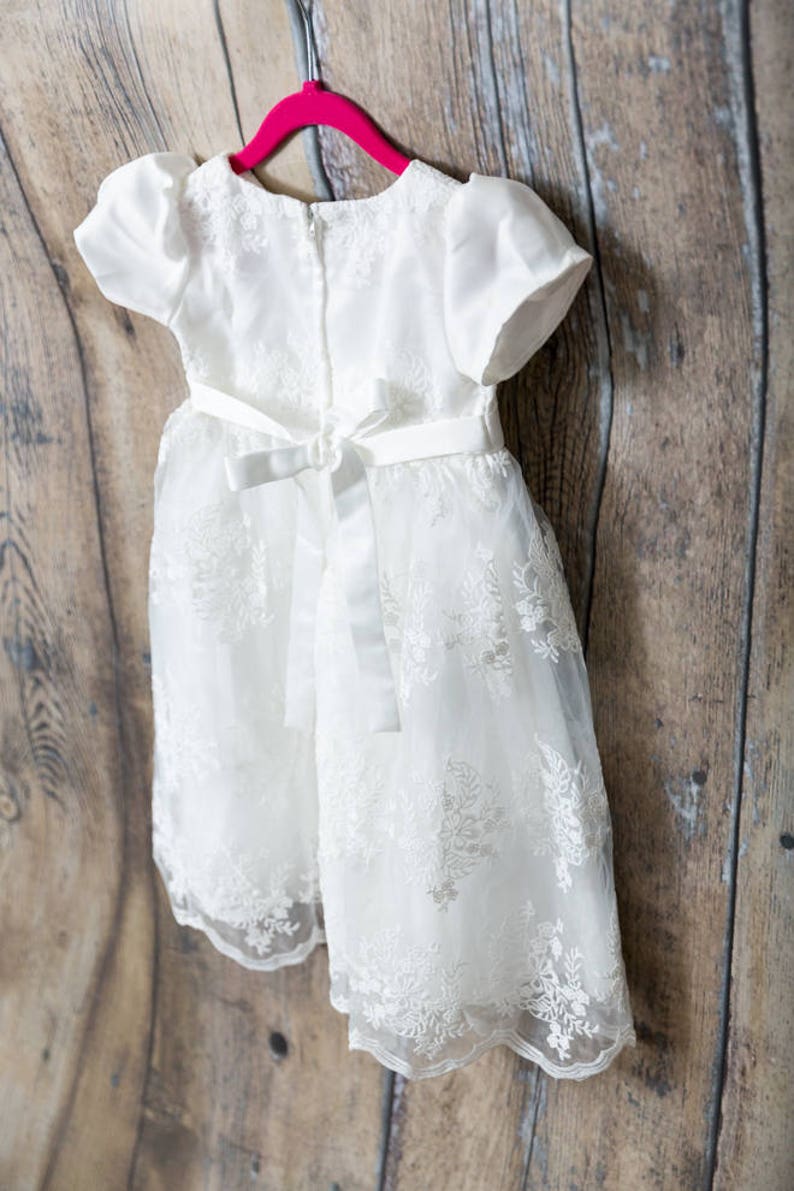 May include: A white dress featuring short puff sleeves, a round neckline, and a back zipper closure. The bodice and skirt are adorned with intricate white floral embroidery over a sheer overlay, finished with a satin ribbon tie at the waist.