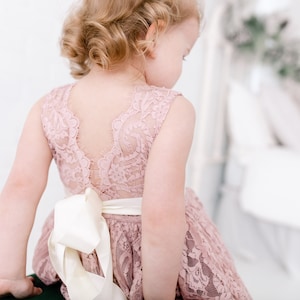 Pu&ograve; includere: Una bambina che indossa un abito di pizzo rosa polvere con un fiocco di raso bianco in vita. L'abito ha una schiena in pizzo.
