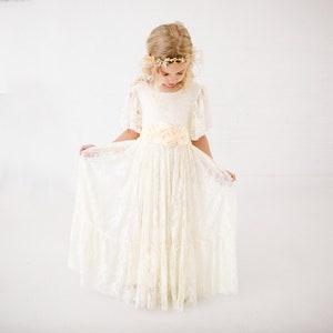 以下が含まれることがあります： 花冠と花柄のサッシュベルトをつけた白いレースのドレスを着た少女。ドレスはフリルスカートと半袖です。
