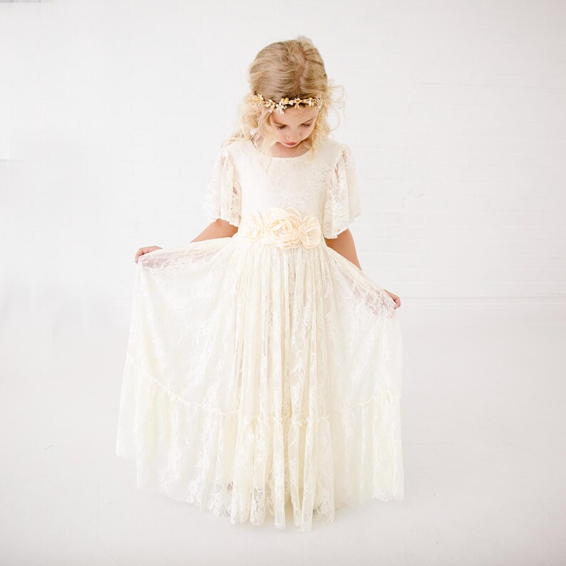 以下が含まれることがあります： 花冠と花柄のサッシュベルトをつけた白いレースのドレスを着た少女。ドレスはフリルスカートと半袖です。