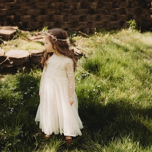 K&ouml;nnte beinhalten: Ein junges M&auml;dchen in einem wei&szlig;en Spitzenkleid und einer goldenen Krone steht in einem grasbewachsenen Bereich. Sie schaut nach links aus dem Bild.