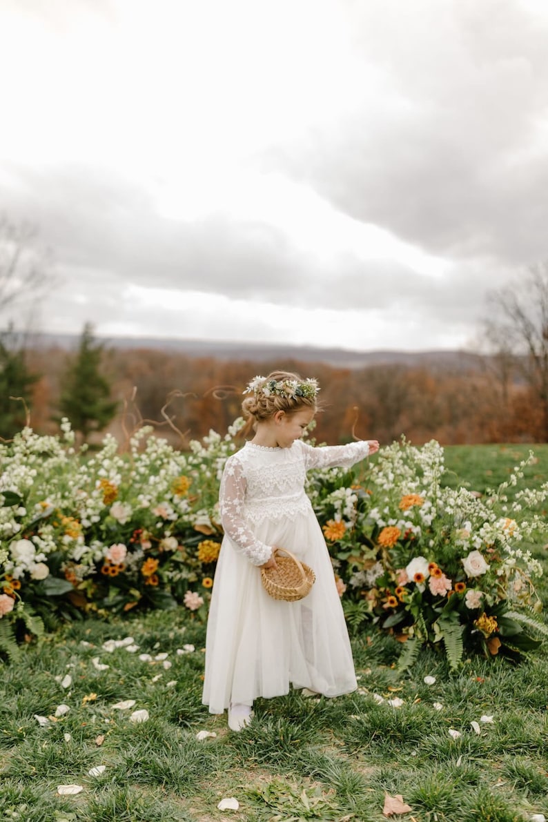 May include: A young girl in a white lace dress and flower crown walks down a flower-covered aisle, holding a small wicker basket. She is surrounded by green grass and white flower petals.