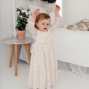 May include: A young child wearing a white lace dress with a flower embellishment on the waist. The child is standing in front of a white bed with a white blanket and a furry throw.