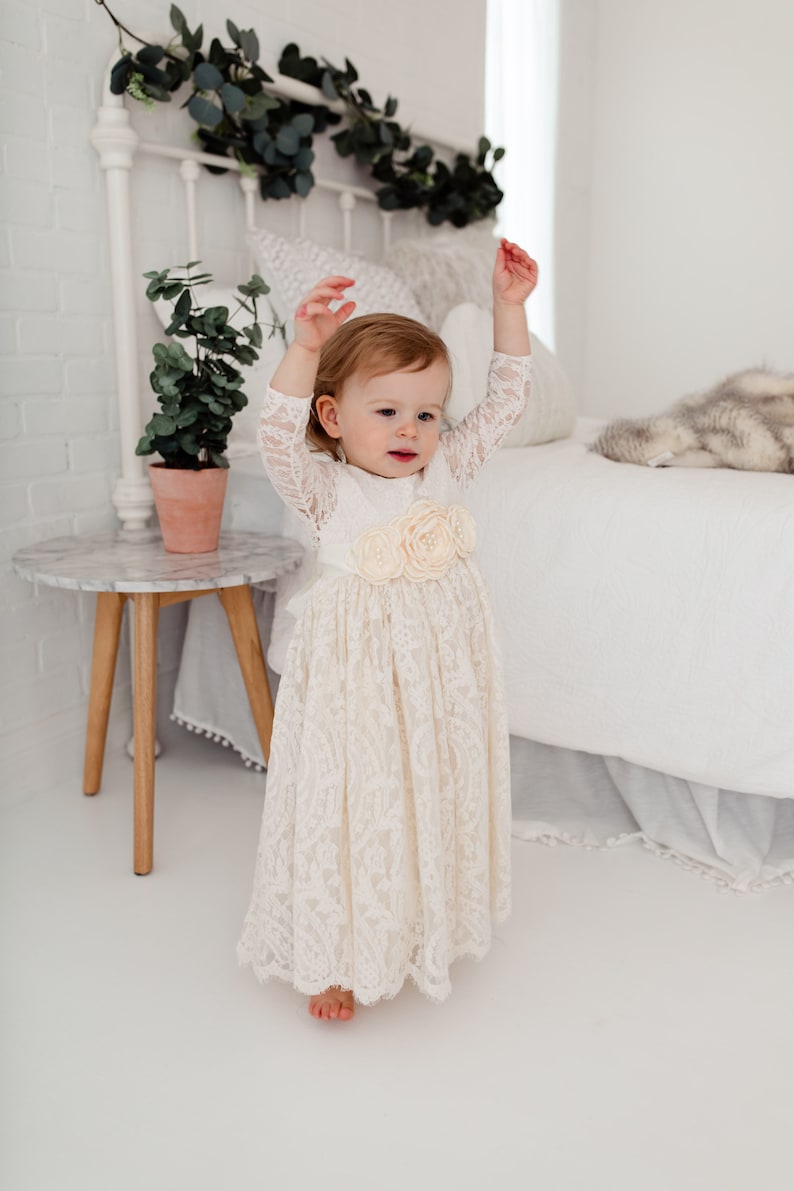 May include: A young child wearing a white lace dress with a flower embellishment on the waist. The child is standing in front of a white bed with a white blanket and a furry throw.