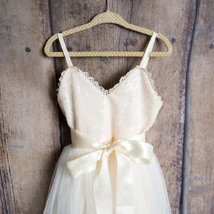 May include: A white tulle dress with a lace bodice and a satin sash. The dress is hanging on a wooden hanger against a woodgrain background.