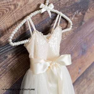 May include: A white tulle dress with a sequined bodice and a satin sash tied in a bow. The dress is hanging on a pearl-beaded hanger.